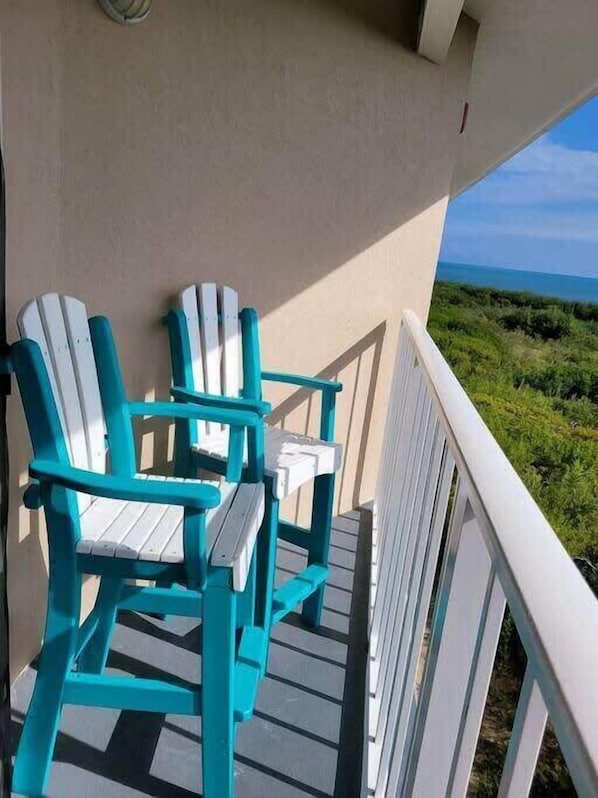 A balcony with two turquoise Adirondack chairs and a small table, overlooking a coastal landscape.