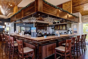 A bar area with a wooden bar counter, multiple stools, and a ceiling with wooden beams and greenery.