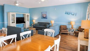 A room with a wooden dining table, blue sofas, a TV, and a ceiling fan. - Spacious 6BR Oceanview Condo w/ Pool & Hot Tub (North Myrtle Beach)