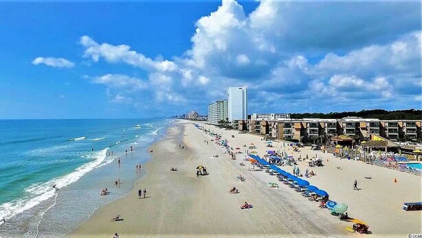 A beach with a long stretch of sand, umbrellas, and beachgoers. - Spacious 6BR Oceanview Condo w/ Pool & Hot Tub (North Myrtle Beach)