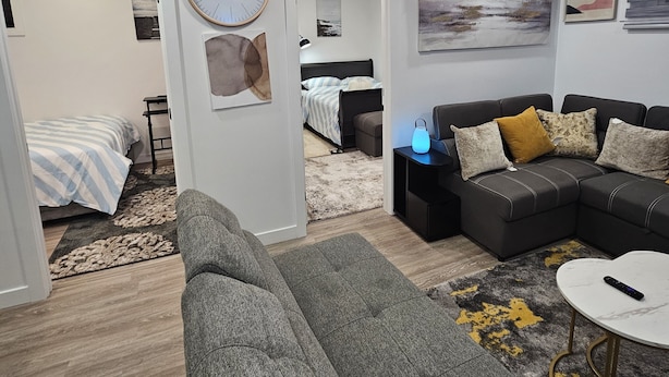 Un salon moderne avec un canapé gris, une petite table basse et une table d’appoint noire sur laquelle se trouve une lampe. On aperçoit, au fond, un lit recouvert d’une couverture à rayures.