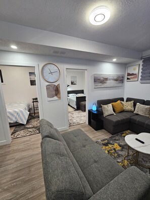 A modern living room with a grey sofa, a small coffee table, and a black side table with a lamp. There is a bed with a striped blanket in the background.