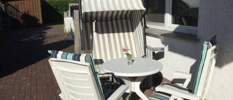 Een terras met twee witte stoelen en een kleine tafel, een gestreept zonnescherm en een huis op de achtergrond.