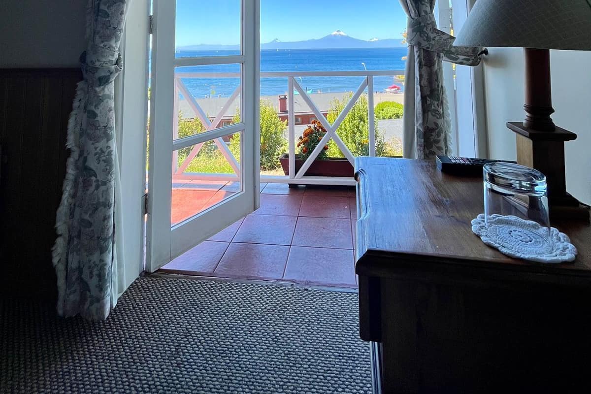Una habitación con vistas al mar y a las montañas, una mesa de madera con una lámpara, y una puerta que da al exterior.