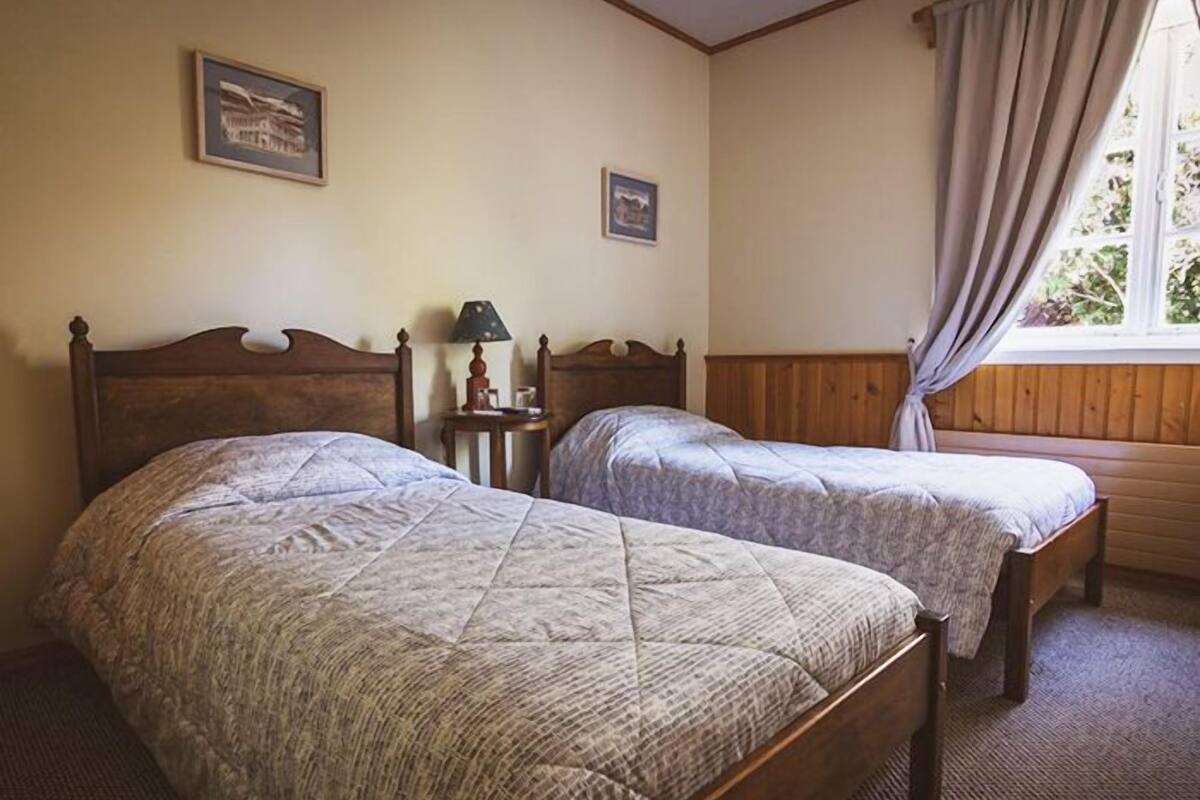 Una habitación con dos camas individuales, cabeceras de madera, una mesita de noche con lámpara y cuadros en la pared.