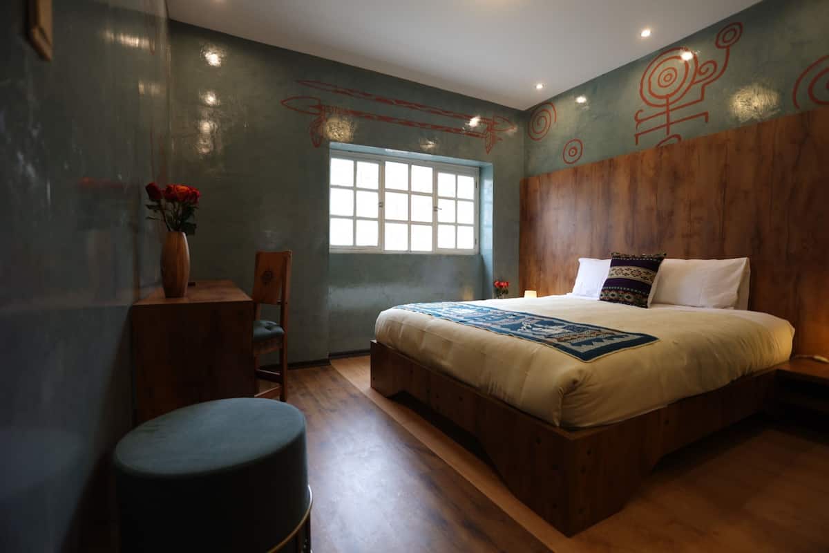 Une chambre avec une tête de lit en bois, un grand lit, une fenêtre, un bureau avec une chaise et un vase rempli de fleurs.
