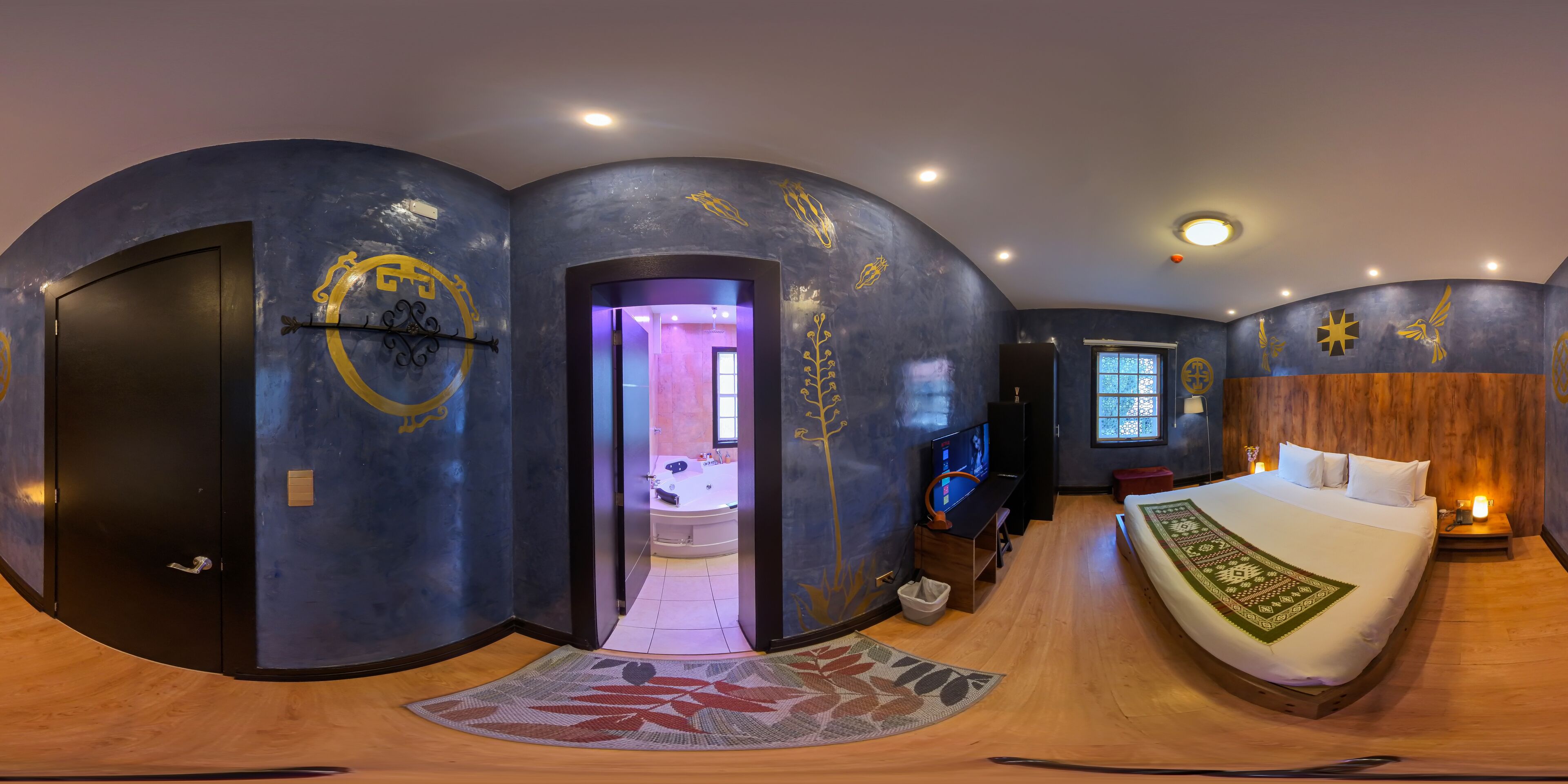 Una habitación de hotel moderna con un diseño de pared curva, una cama grande con colcha estampada, un televisor y una mesita de noche con lámpara.