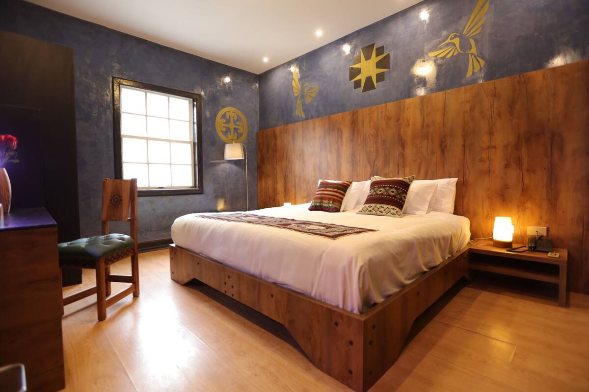 Une chambre à coucher avec une tête de lit en bois, un grand lit avec de nombreux oreillers, une table de chevet avec une lampe et une fenêtre avec des stores.