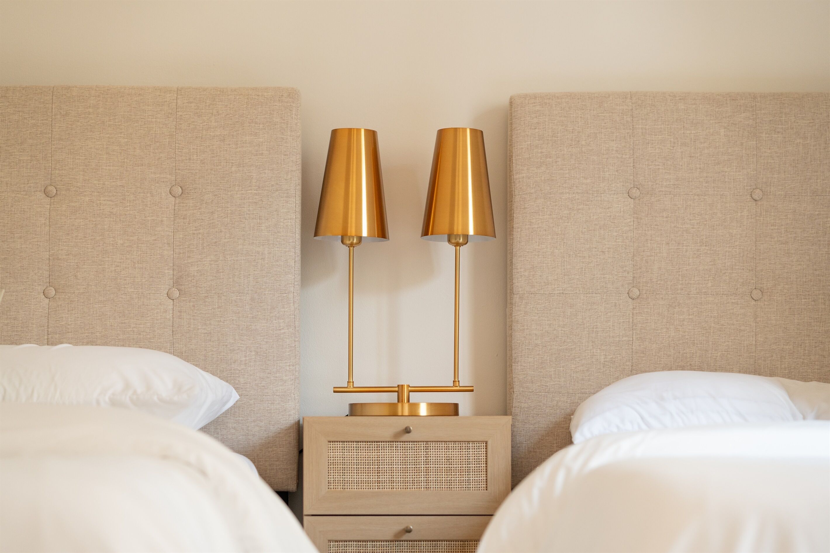 Un lit bien fait, avec du linge de lit blanc, une tête de lit recouverte d’un tissu beige et une table de chevet agrémentée d’une lampe dorée et d’un petit tiroir.