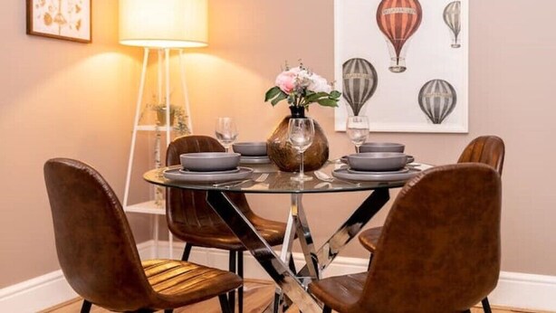 Un coin repas comprenant une table ronde en verre, des chaises à revêtement marron, un vase avec des fleurs et une œuvre d’art murale représentant des montgolfières.