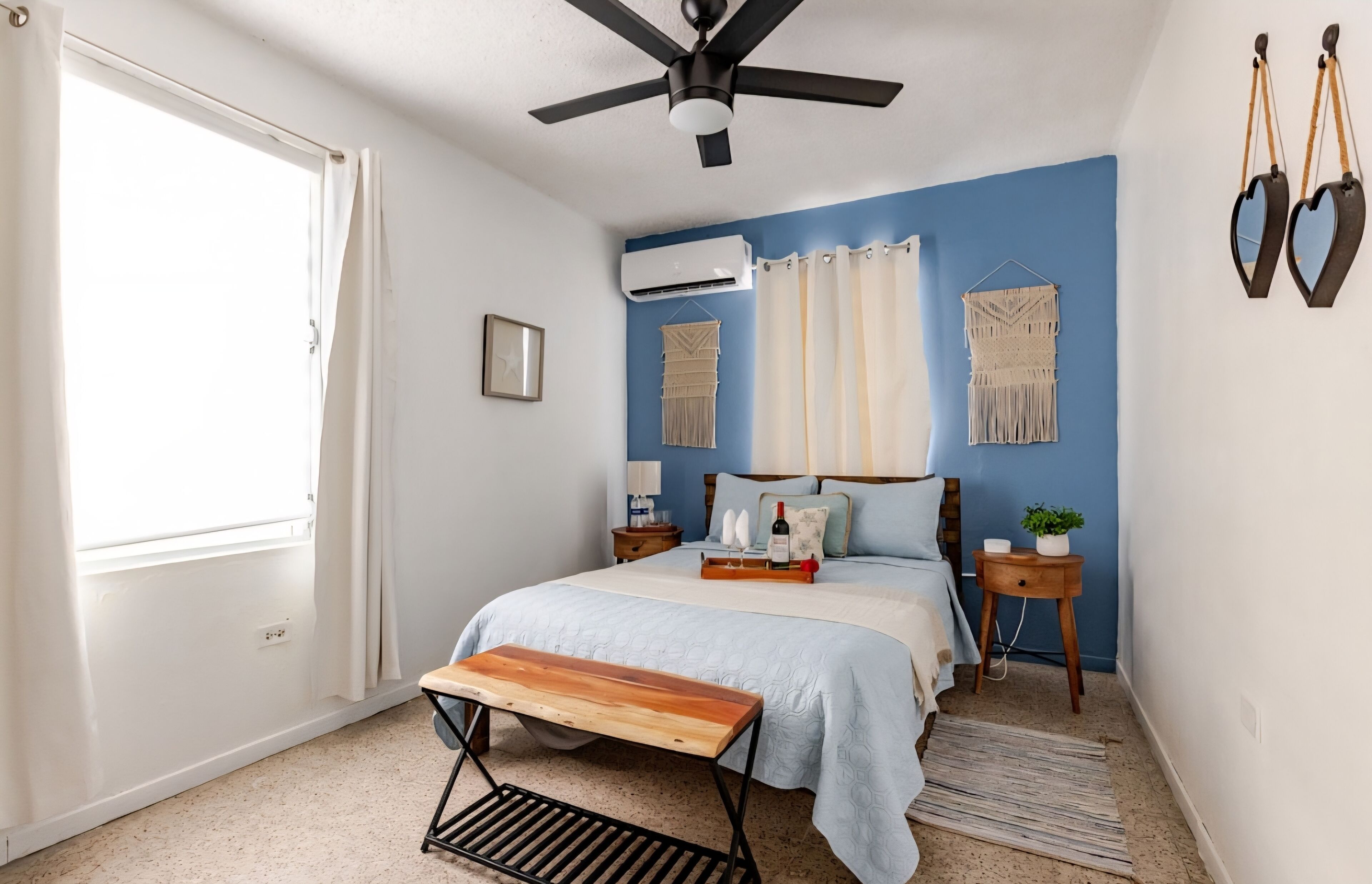 A bedroom with a bed, bedside tables, a bench, a ceiling fan, and a window with curtains.