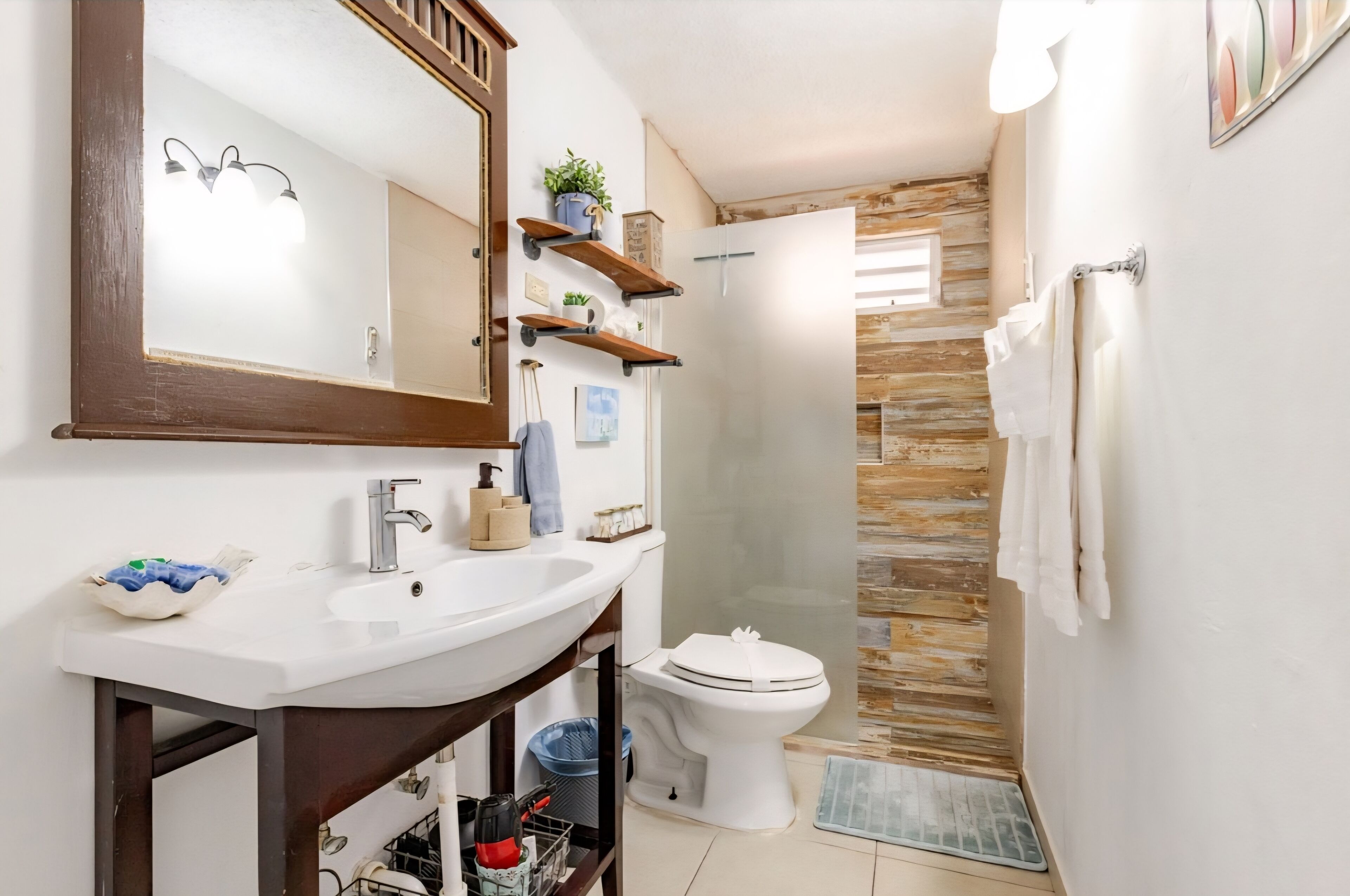 Un baño con un lavabo blanco, un espejo y un inodoro. Hay estantes de madera sobre el lavabo y una zona de ducha con puerta de cristal.