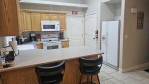 A compact kitchen with wooden cabinets, a white microwave, a stove, and a refrigerator. There is a small island with two stools and a sink. - Vista Rental 1 Cozy Home, 25 min from Zion National Park (La Verkin)