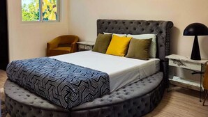 Ein modernes Schlafzimmer mit einem runden Bett, einem gesteppten Kopfteil und einem großen Fenster mit Blick auf Palmen.