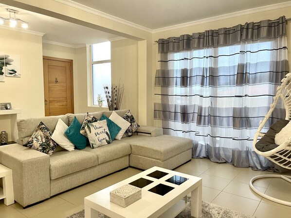 A living room with a beige sectional sofa, a white coffee table, a wicker chair, and a window with grey curtains.