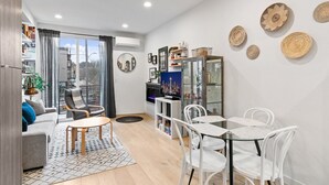 A modern living room with a sofa, coffee table, and dining area with chairs. There is a large window with curtains, a bookshelf, and decorative wall art. - Ravenna Trailside Retreat Walk to UW and U-Village (Seattle)