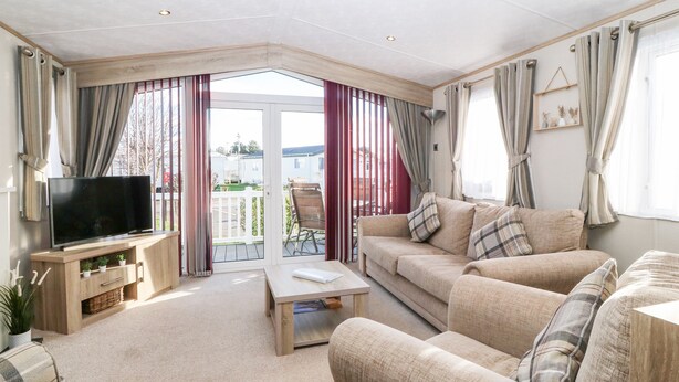A living room with a TV, sofa, and coffee table. There is a balcony with curtains and a view of the outside. - Snape Lodge (Abergele)