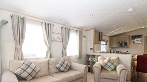 A living room with a beige sofa, a matching armchair, and a coffee table. There is a kitchen area in the background with a microwave and a sink. - Snape Lodge (Abergele)