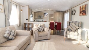 A spacious living room with a sofa, armchair, and a coffee table. There is a dining area with a table and chairs. The room has a kitchen area with cabinets and a microwave. - Snape Lodge (Abergele)