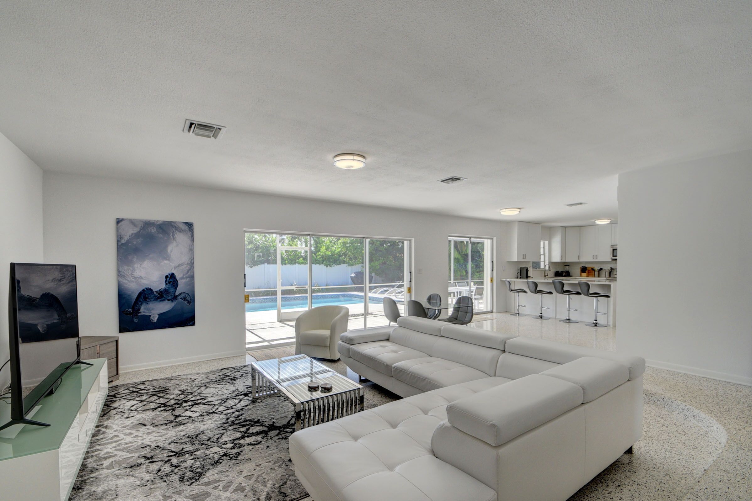 A spacious living room with a large white sofa, a glass coffee table, and a view of a swimming pool through large windows.