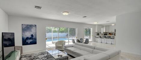 A spacious living room with a large white sofa, a glass coffee table, and a view of a swimming pool through large windows.