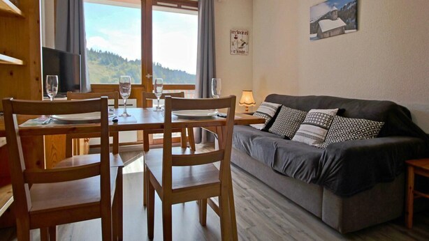 A cozy room with a dining table set for four, a sofa with patterned cushions, a TV on a wooden stand, and a scenic view through the windows. - 10m from ski slopes, 1st floor, balcony, tv, 34m², Chamrousse (Chamrousse)