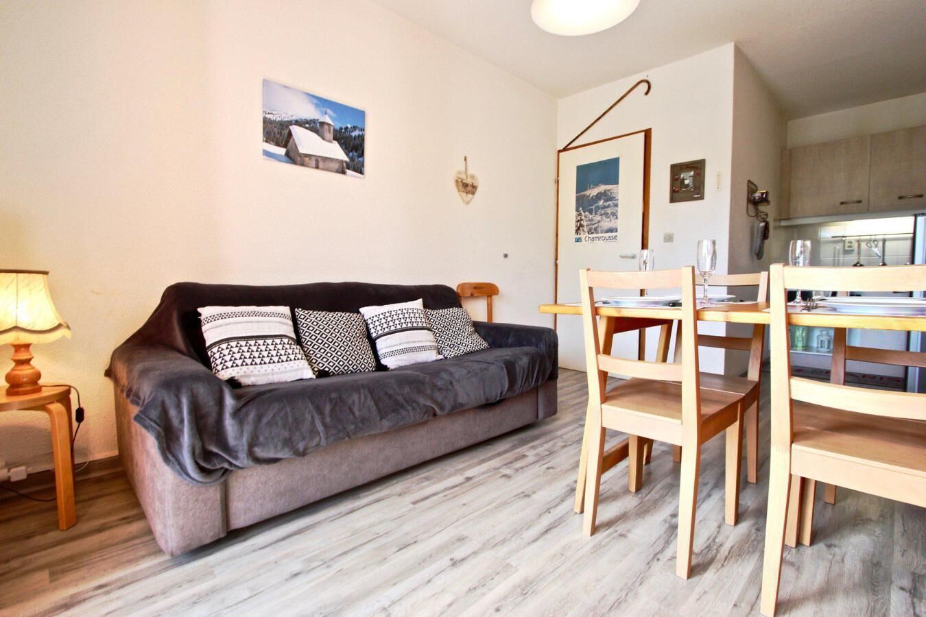 Un salon chaleureux avec un canapé gris, des coussins à motifs, une table à manger en bois avec des chaises, et une cuisine en arrière-plan.