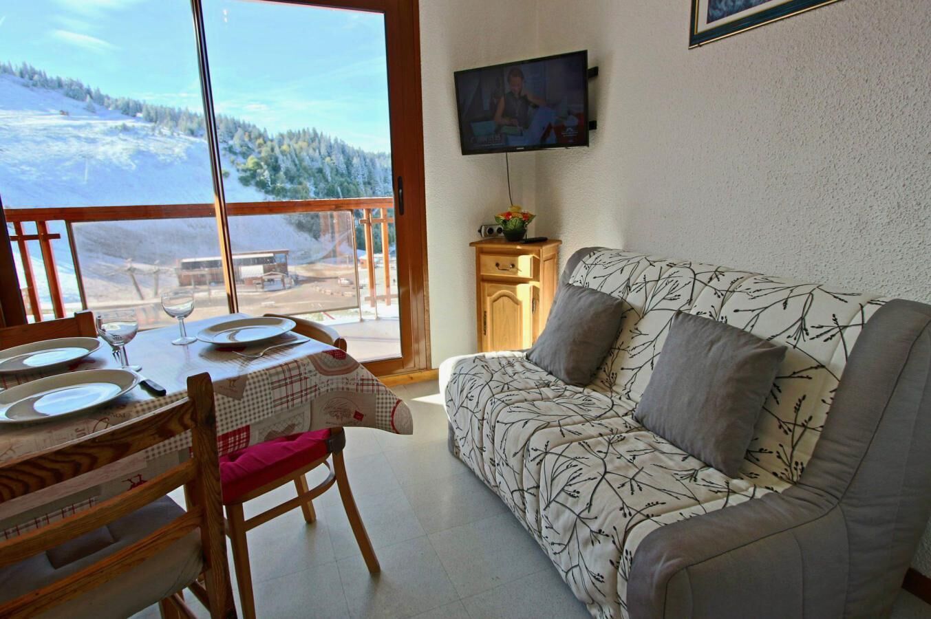 Een gezellige kamer met een bank en een eettafel gedekt voor twee personen, een televisie aan de muur, en zicht op een besneeerd landschap door grote ramen.