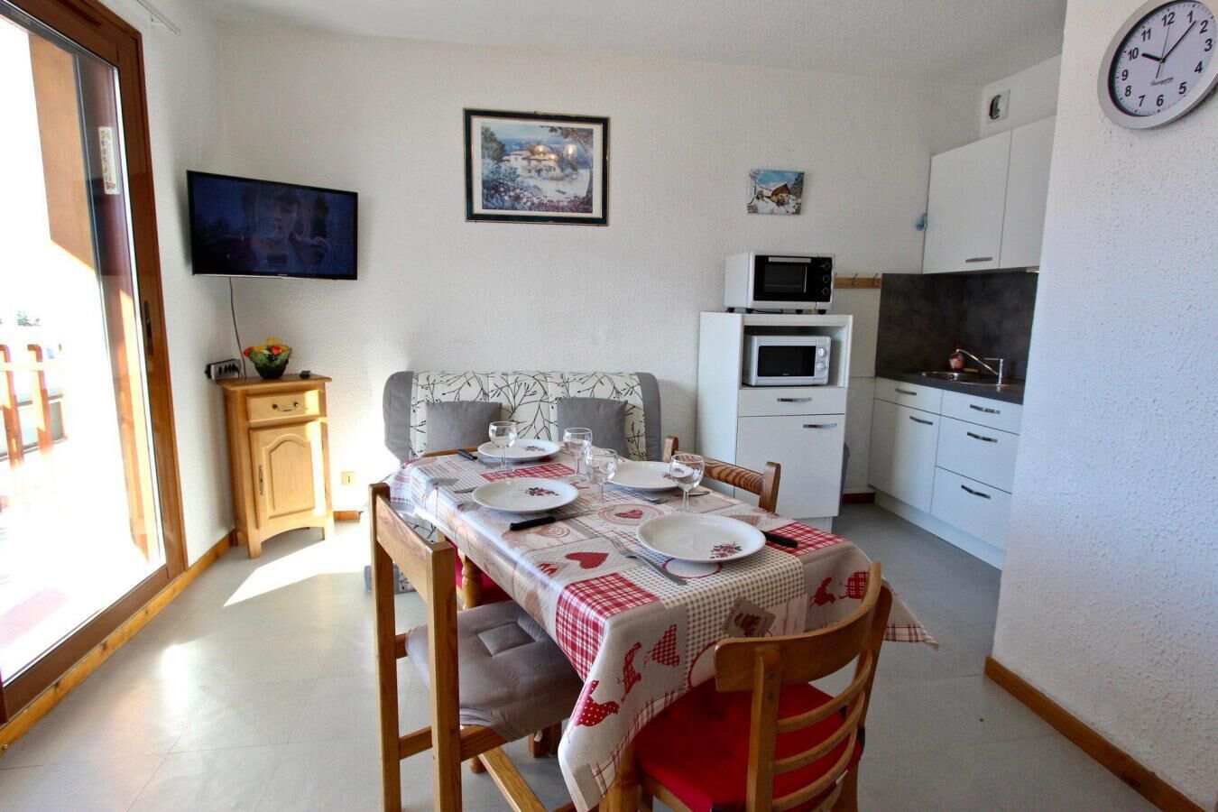 A dining area with a table set for four, a television on the wall, a microwave, and a clock.