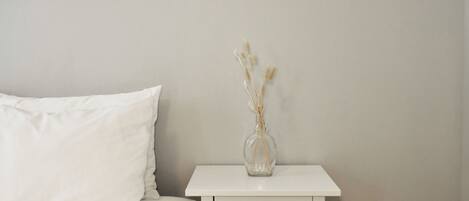 A bed with white bedding, a white nightstand with a drawer, and a clear vase with dried plants on the nightstand.