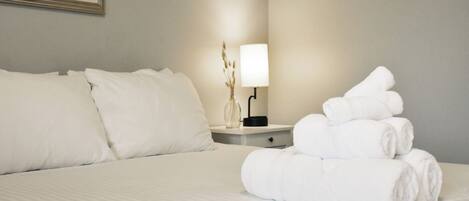 A neatly made bed with white linens and pillows, a bedside lamp, and a stack of rolled towels on the nightstand.