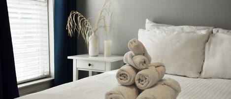 Una cama bien tendida con toallas enrolladas, una mesita de noche con un florero y una vela, y una ventana con persianas.