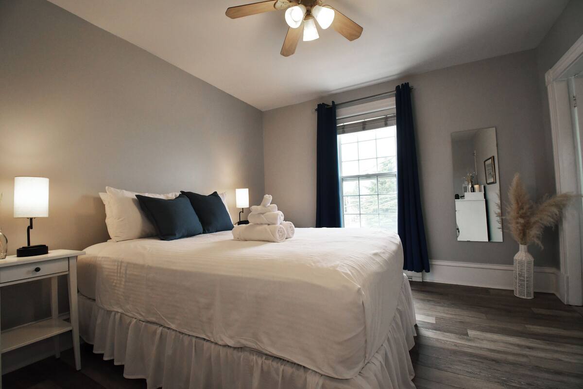 A neatly made bed with white linens and dark pillows, a bedside table with a lamp, a window with curtains, and a mirror on the wall.