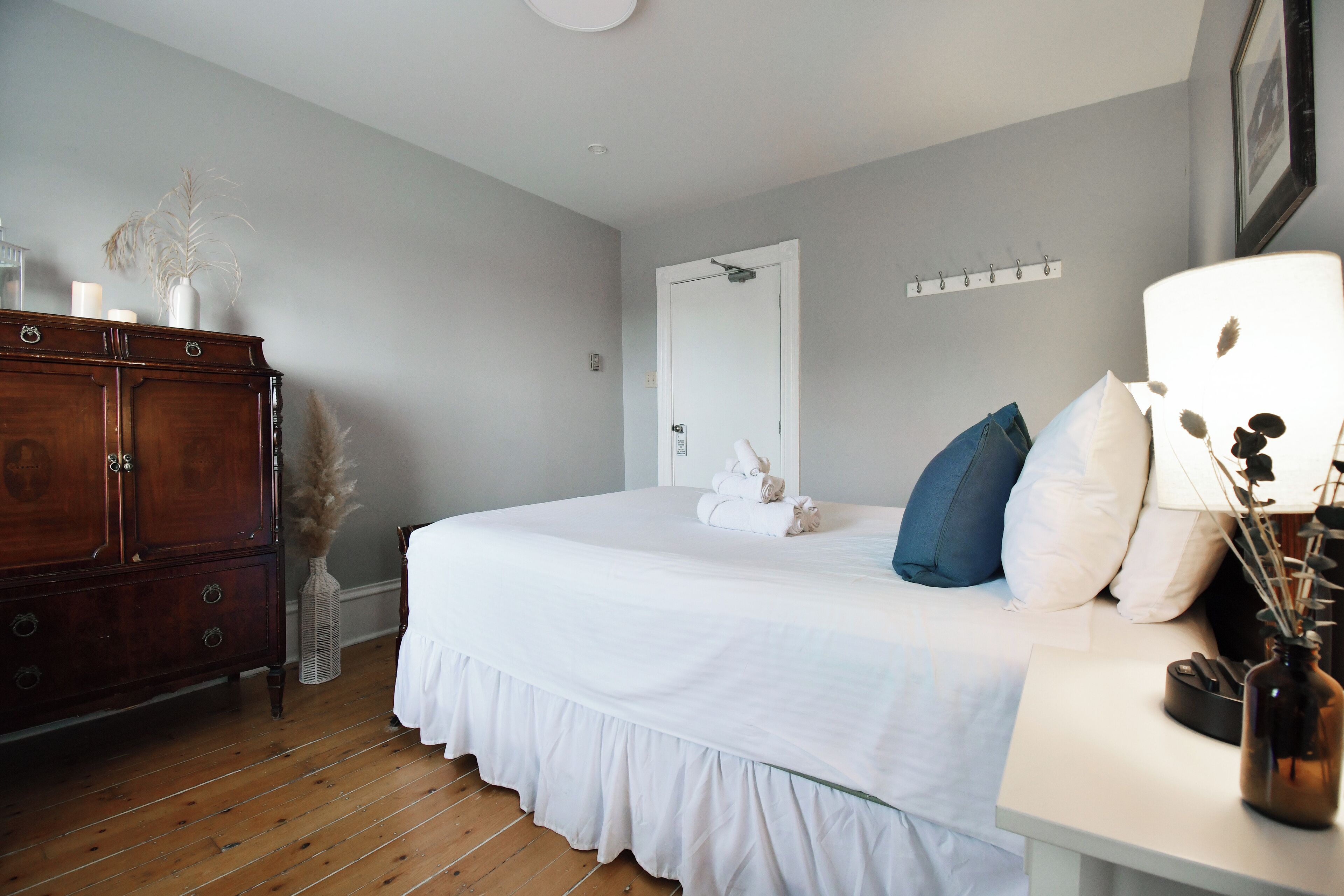 A bedroom with a wooden dresser, a bed with white bedding, and a bedside table with a lamp and vase.