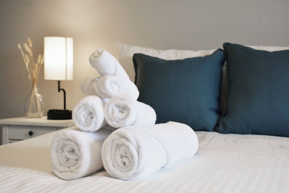 A neatly made bed with rolled white towels, a bedside lamp, and dark green pillows.