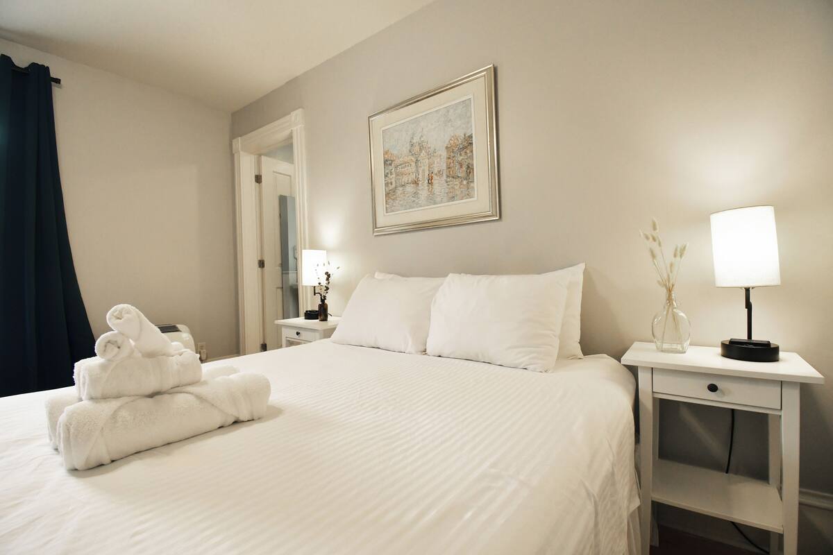 A neatly made bed with white linens, a framed painting on the wall, and a bedside table with a lamp and a vase.