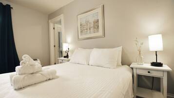 A neatly made bed with white linens, a framed painting on the wall, and a bedside table with a lamp and a vase.