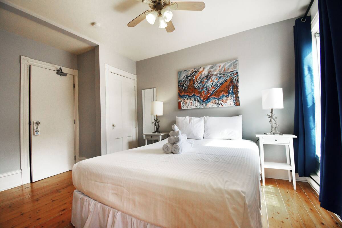 A hotel room with a large bed, a ceiling fan, a painting on the wall, and a window with curtains.
