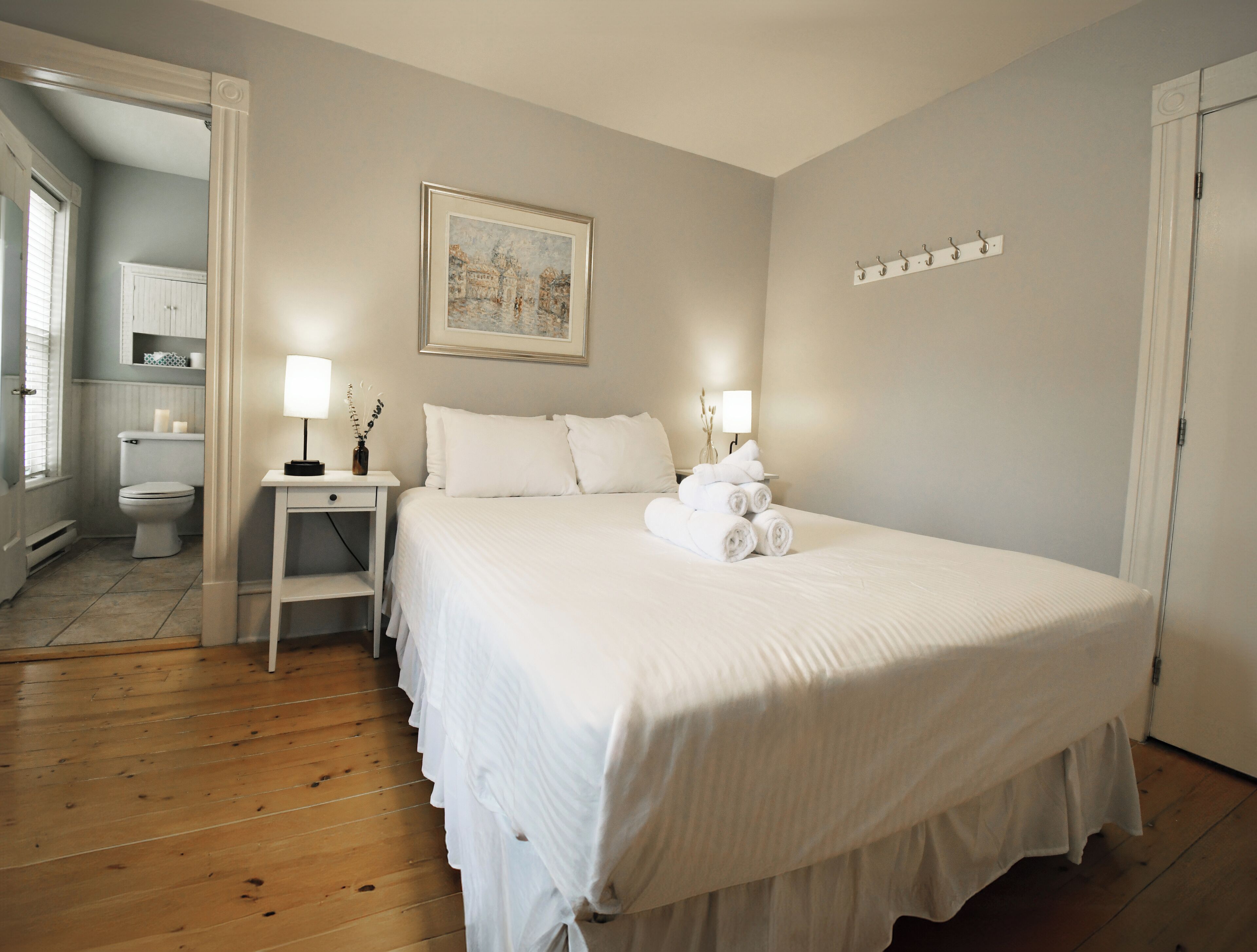Ein ordentlich eingerichtetes Schlafzimmer mit einem Bett, Nachttischlampen, einem gerahmten Bild an der Wand und einem durch eine geöffnete Tür einsehbaren Badezimmer.