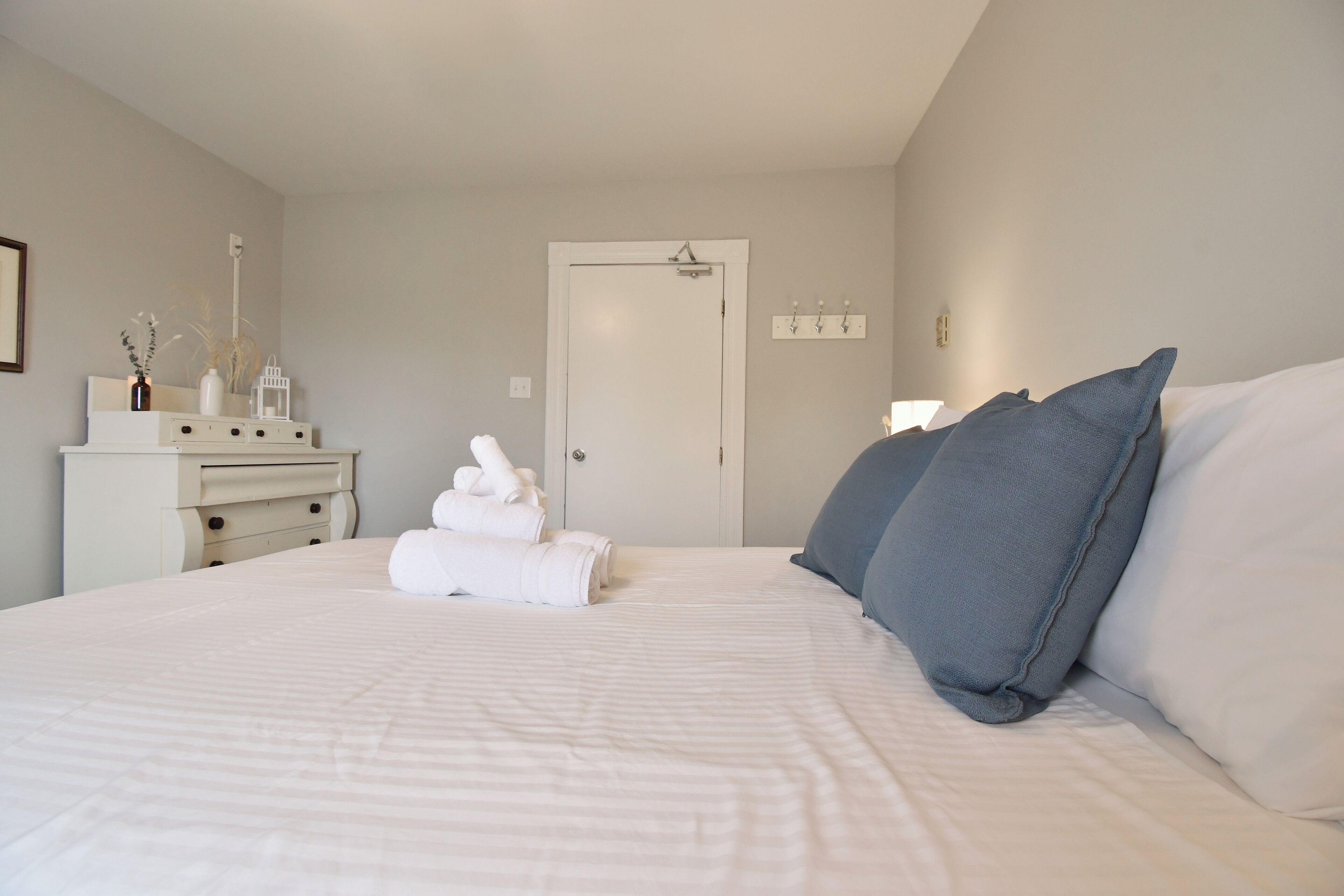 Una cama bien tendida con sábanas blancas y almohadas azules, un tocador blanco con un jarrón y una lámpara, y una puerta con interruptor de luz y ganchos en la pared.
