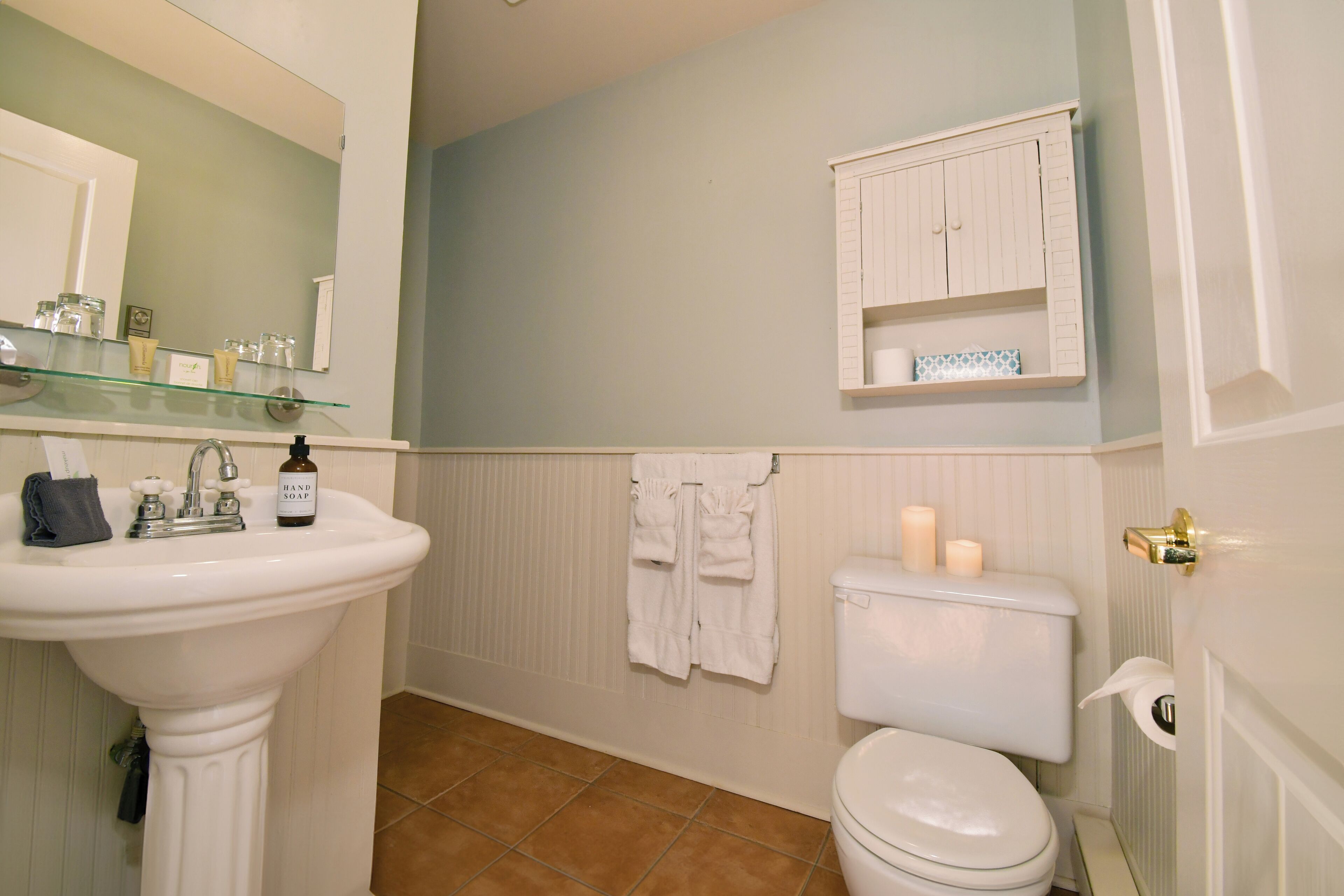 Une salle de bain avec un lavabo sur colonne, des toilettes, une étagère en verre contenant des articles de toilette et un meuble blanc au-dessus des toilettes.