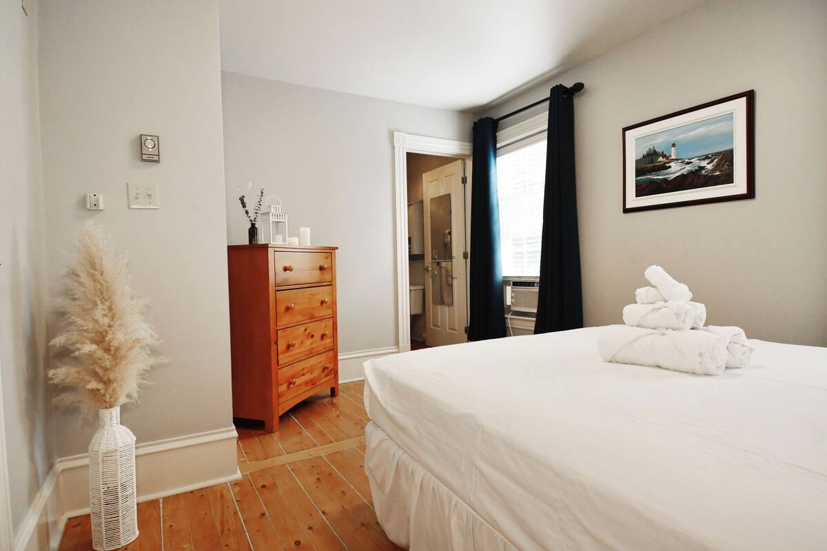 A bedroom with a wooden dresser, a white bed with a white comforter, a vase with pampas grass, a framed picture on the wall, and a view of the bathroom through an open door.