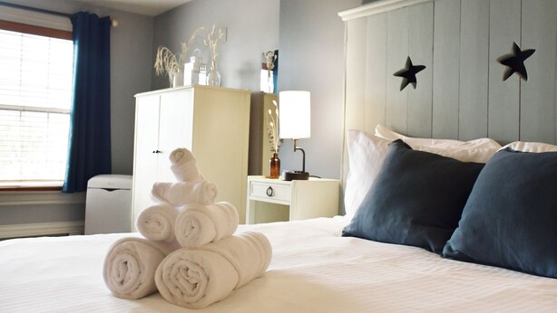 A neatly made bed with rolled towels, a nightstand with a lamp, a dresser with decorative items, and a wall with star decorations. - Hotel Gabriel by Bower Boutique Hotels (Shediac)