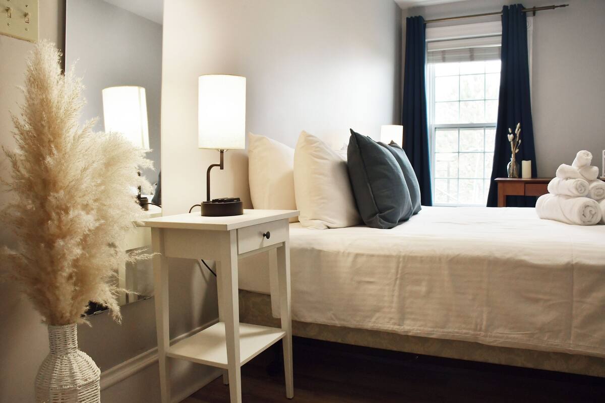 A neatly made bed with white and dark pillows, a bedside table with a lamp, and a vase with dried plants.