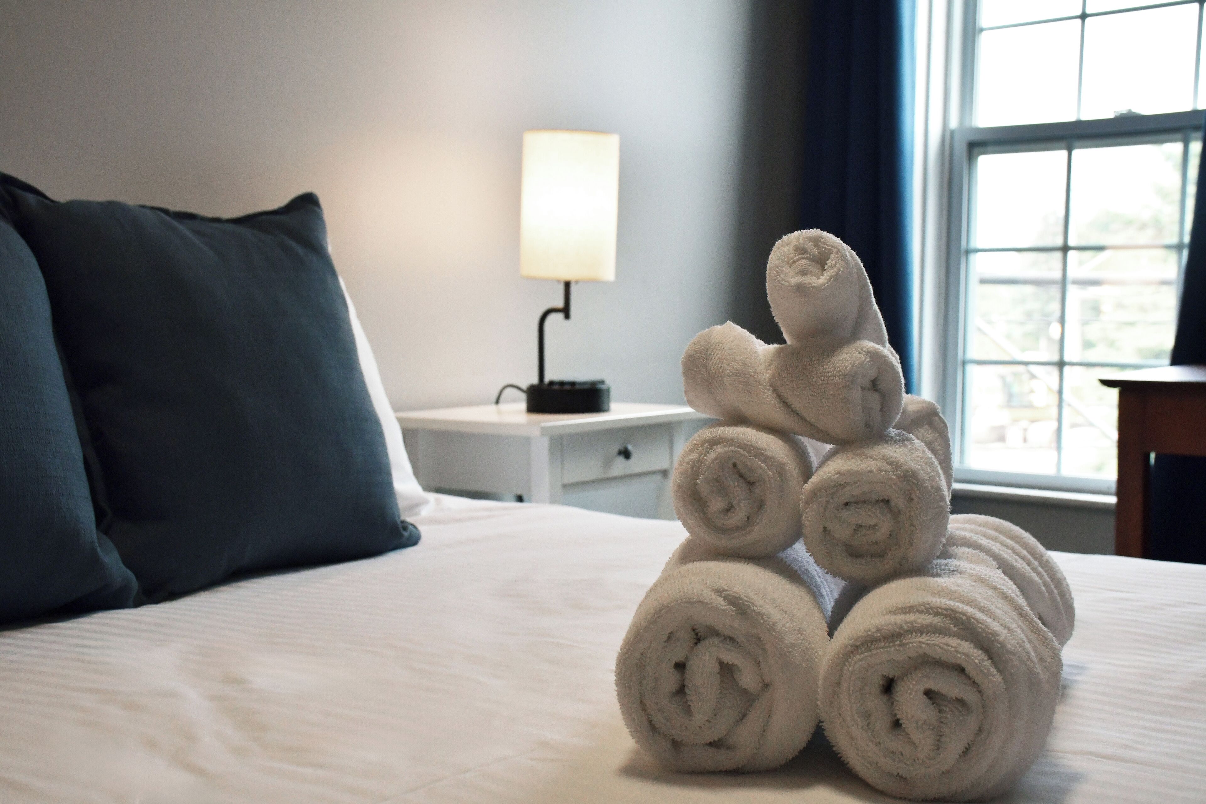 A neatly made bed with rolled towels stacked in a pyramid shape, a bedside lamp, and a window with curtains.