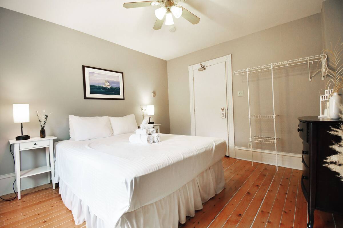 A bedroom with a large bed, a bedside table with a lamp, a framed picture on the wall, a wardrobe, and a ceiling fan.