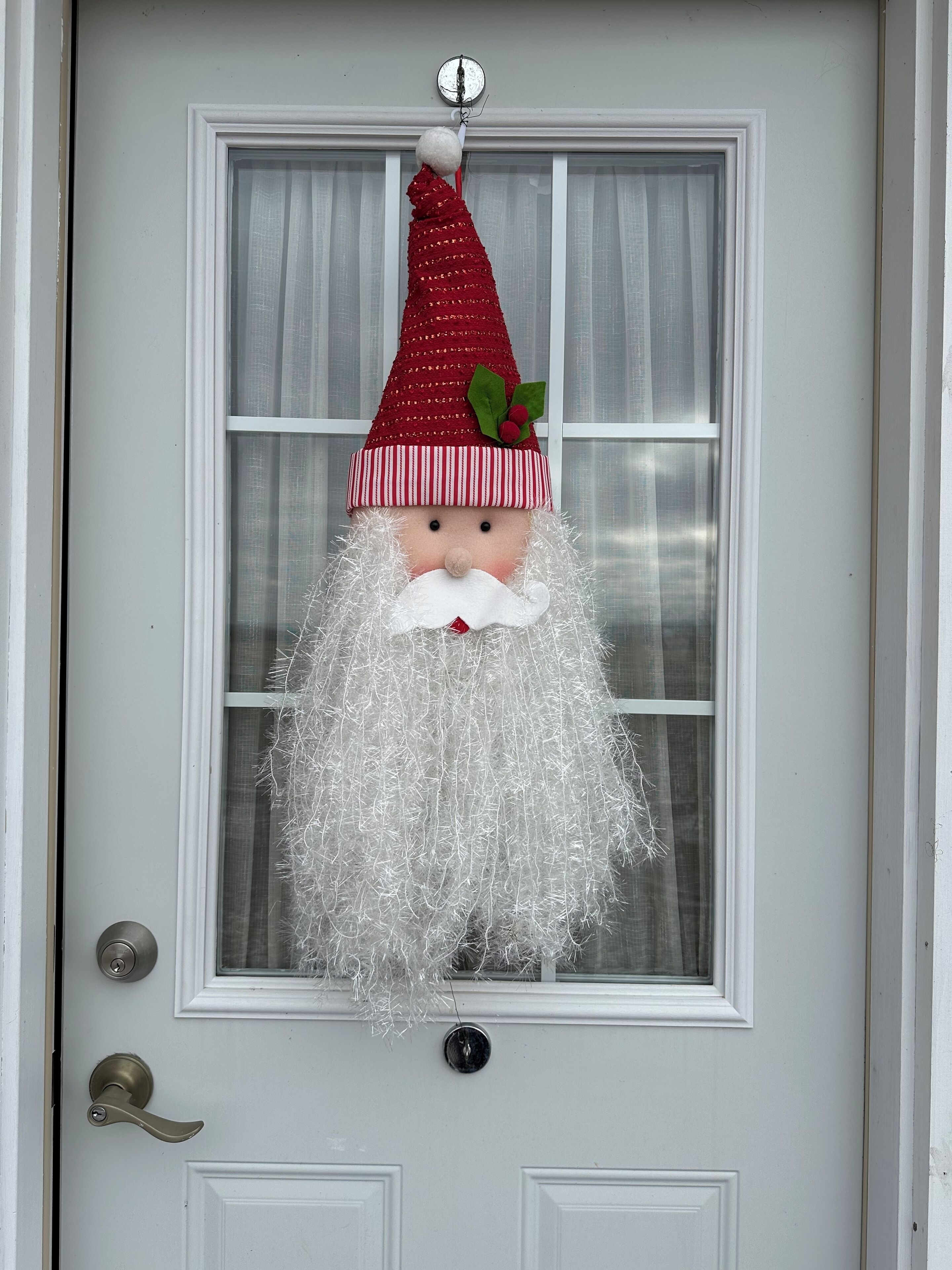 Una porta con un decorativo ornamento natalizio raffigurante Babbo Natale appeso sopra una finestra con tende bianche.