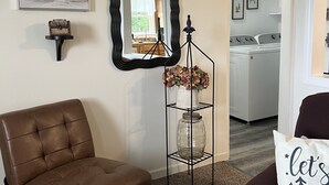 Un salon avec un fauteuil en cuir marron, un miroir à cadre noir, un vase de fleurs et un coussin blanc orné d’un motif d’arbre.