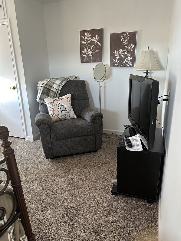 A living room with a grey armchair, a television on a stand, a floor lamp, and two wall-mounted floral artwork pieces.