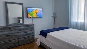 Une chambre à coucher avec un lit, une commode, un téléviseur fixé au mur et une fenêtre avec des rideaux.
